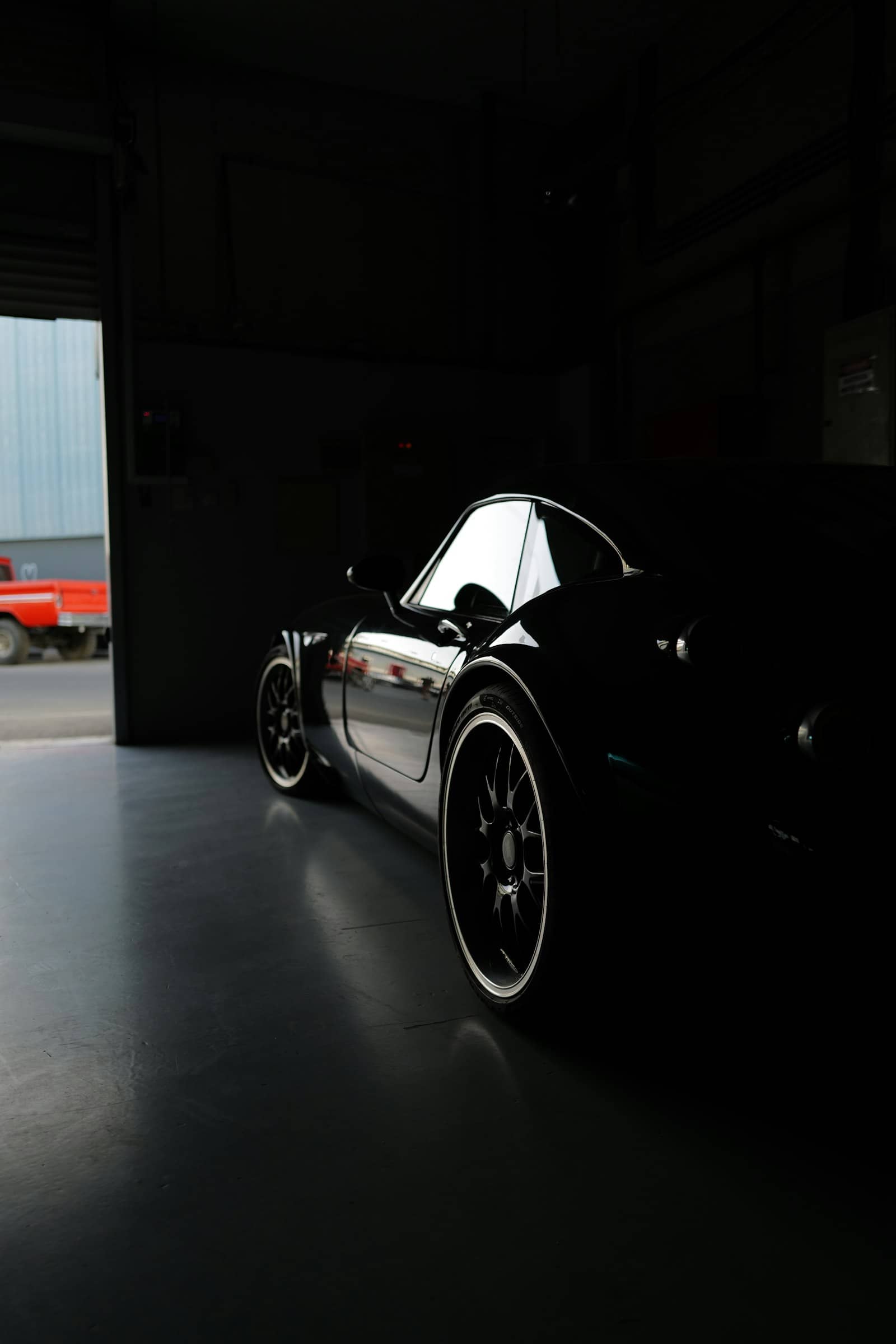 Black coupe alone in the dark studio bay, gloss catching the light from the open door